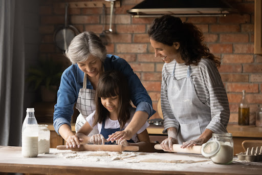A grandmother, young grandaughter and mother bake together at a large butcher block table. Gifting stocks can benefit both the giver and the recipient.