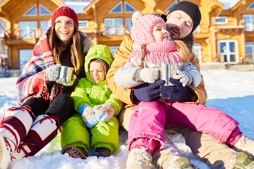 A family with two young children sitting in the snow drinking hot chocolate in front of their investment real estate property.