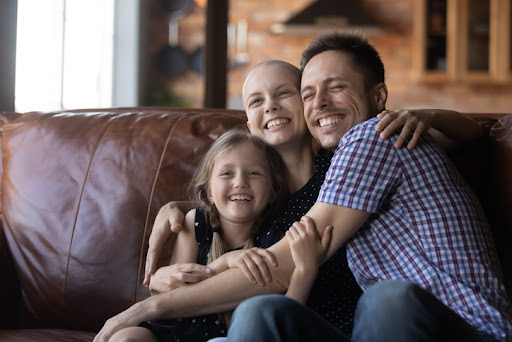 A family sitting on a leather couch smiling and hugging a young mom with a terminal illness.