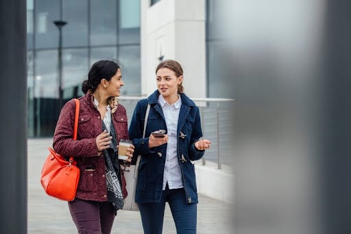 Businesswomen are walking through the city. They are talking and one is holding a coffee while the other is holding a smartphone. Answering questions about asset allocation.