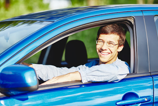 A teenager smiling from the driver’s seat window in a blue car. New driver car insurance.